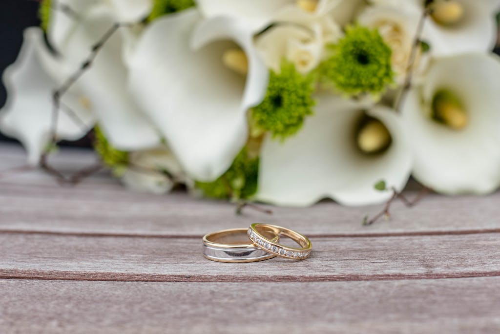 Home Close-up of wedding rings on wooden surface with white and green floral arrangement.