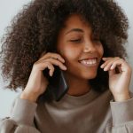 Home Smiling woman with afro hair enjoying a phone call indoors, wearing a casual sweater.